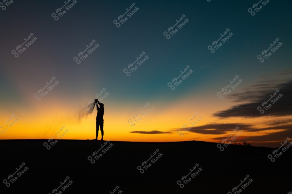 Silhouette of a person standing on sand dunes during sunset, raising sand in the air with their hands. The sky transitions in colors from deep blue to bright orange with sunset gradients in the background.