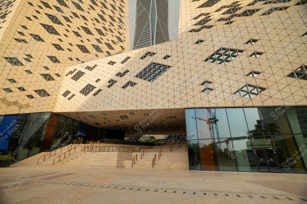 Image of a modern building with a unique architectural design, featuring an interlocking triangular facade with slanted angles and small windows in various shapes scattered irregularly. The building is shown in daylight with a clear sky in the background. The entrance features a grand staircase with metal railings, and transparent glass facades are visible on the sides.