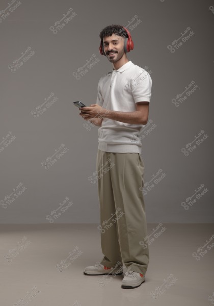 A portrait of a young man wearing red headphones, a white polo shirt, and beige pants, standing and holding a phone in his hands. The man is smiling and looking at the camera, with a simple gray background.
