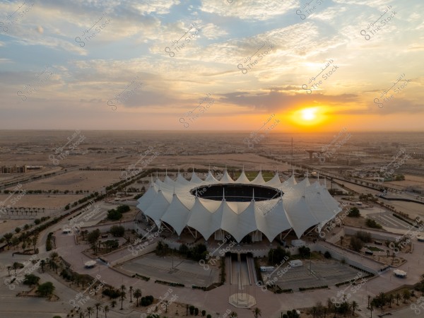 Aerial view of a uniquely designed sports stadium in the middle of a desert, surrounded by palm trees and roads, with a stunning sunset in the background. The sky is filled with illuminated clouds, and the horizon stretches towards a distant city, creating an enchanting scene.