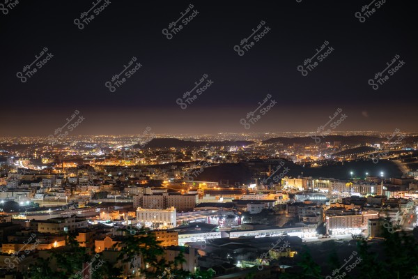 A nighttime cityscape illuminated with streetlights and building lights, creating a striking contrast against the dark night sky. Urban sprawl extends to the horizon, with low hills visible in the background. The lighting highlights various architectural designs of the buildings.