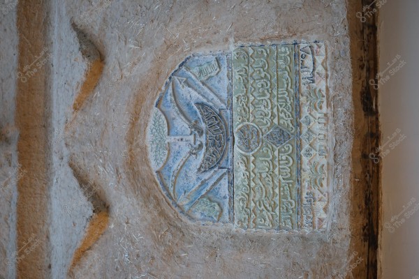 A multicolored wall plaque featuring Arabic inscriptions and decorations. The plaque appears to be part of an ancient wall, showcasing architectural details and colorful botanical motifs.