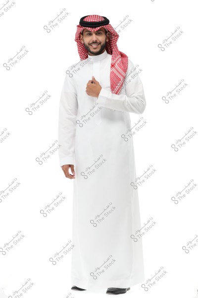 Image of a man wearing traditional Saudi attire, consisting of a white thobe, red checkered ghutra, and a black agal. The man is smiling slightly and stands against a white background.