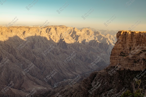 A landscape image featuring rocky mountains with sharp terrain, appearing in shades of brown. The mountain in the foreground casts a dark shadow, while the background features a range of mountains bathed in golden sunlight, under a clear sky above.
