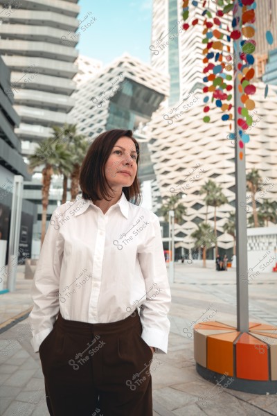 A photo of a woman wearing a white shirt and brown pants, standing in a modern urban environment with contemporary architectural buildings in the background. There are palm trees and colorful decorations hanging. The sky is blue in the background.
