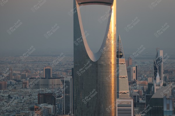 صورة لمدينة الرياض في المملكة العربية السعودية تُظهر برج المملكة البارز بتصميمه الفريد, الذي يأخذ شكل قوس عالٍ. البرج مُضاء بأشعة الشمس, ويظهر بجانبه برج الفيصلية بتصميمه الهرمي المميز وكرته الزجاجية. تحيط المباني الشاهقة الأخرى في الأفق حيث يبدو الغروب في الخلفية.