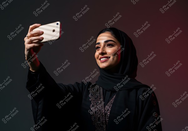 A portrait of a woman wearing a traditional black abaya and hijab, taking a selfie with a smartphone. The background is dark with subtle lighting that highlights her facial features and attire.