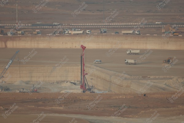 Image of a large construction site in a desert area. The image shows a large red drilling machine surrounded by white trucks and other heavy machinery. Behind the site, there is a highway with several trucks and cars passing along it. The surrounding land is dry and expansive, indicating a major construction process underway in an open area.