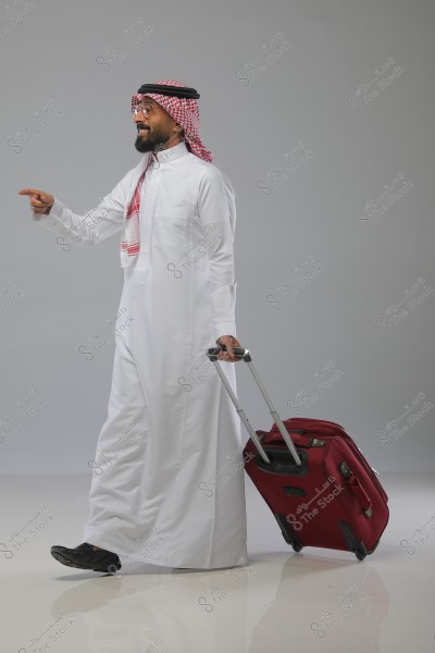 A man wearing a white thobe, a red checkered ghutra, and agal, walking and pulling a red wheeled suitcase. The background is plain gray.