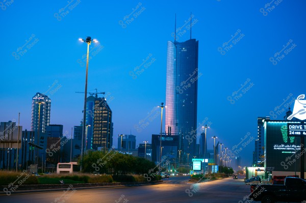 View of modern buildings and skyscrapers illuminated by streetlights at night. A tall glass skyscraper is in the center against a calm blue sky, surrounded by other buildings with contemporary designs and exterior lighting. On the right, a lighted sign advertises a 24-hour service.