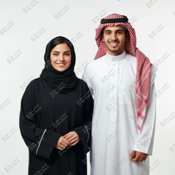 The image shows a man and a woman wearing traditional Saudi attire. The man is dressed in a white thobe and a red and white checkered ghutra with a black agal, while the woman is wearing a black abaya and a black hijab. The background is white, highlighting their details and clothing. Both are smiling and standing next to each other.