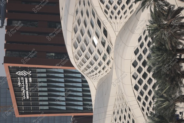 An image of a modern building featuring a glass facade with a geometric mesh design, next to another building displaying the logo "National Development Fund." The building is surrounded by palm trees, reflecting the contemporary architectural style in Saudi Arabia.