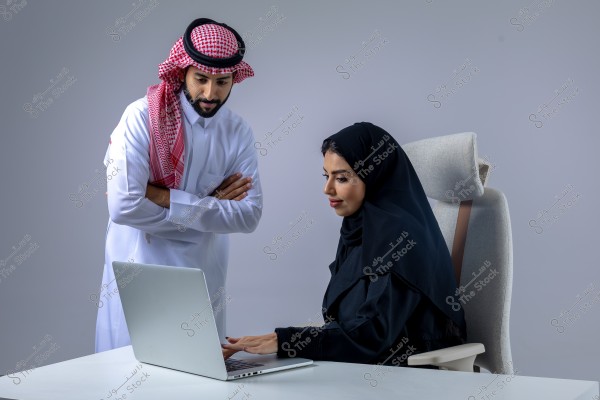Image of a man and a woman working together in an office environment. The woman is seated in a comfortable chair, using a laptop, dressed in a black abaya and hijab. The man is standing beside her, looking at the screen, dressed in a white thobe and a red and white checkered ghutra with an agal, suggesting that his attire might be traditional Saudi clothing.