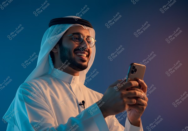 Portrait of a man wearing a white thobe with a traditional headscarf and agal, captured in a moment of happiness while holding a smartphone. The man is smiling, suggesting he is relaxed or finding something intriguing on the phone. The lighting highlights his face against a dark blue background, creating a calm atmosphere.
