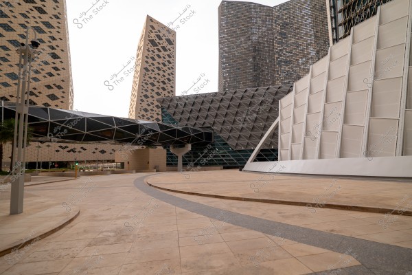 A modern architectural view showcasing a group of buildings with glass facades and unique geometric patterns. The image features an enclosed bridge connecting the buildings and a water panel in the foreground. The towering structures have beige and black slanted designs, and the front building has a 3D metal facade.