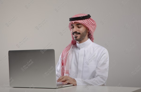 A portrait of a man sitting in front of a laptop, wearing traditional white thobe and a red and white checkered shemagh with an agal. He appears to be working or interacting with the device in a simple office setting. The attire suggests he might be from Saudi Arabia.