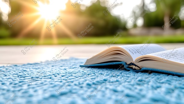 An open book on a blue prayer rug with bright sunlight shining in the background, illuminating the scene. The Arabic script is clearly visible on the book\'s pages, with a blurred green background suggesting a garden or natural area.