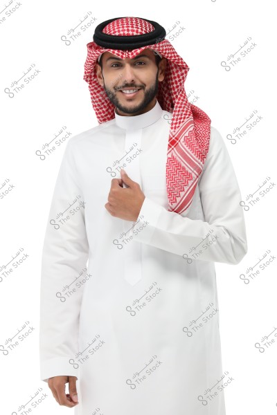 A portrait of a man wearing traditional Saudi attire, featuring a white thobe and a red ghutra with a black agal. The man is smiling against a white background.