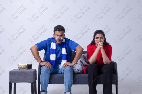 Portrait of a Saudi young man and woman watching a match and cheering their favorite teams cheerfully while having a snack on a blue background, the World Cup.