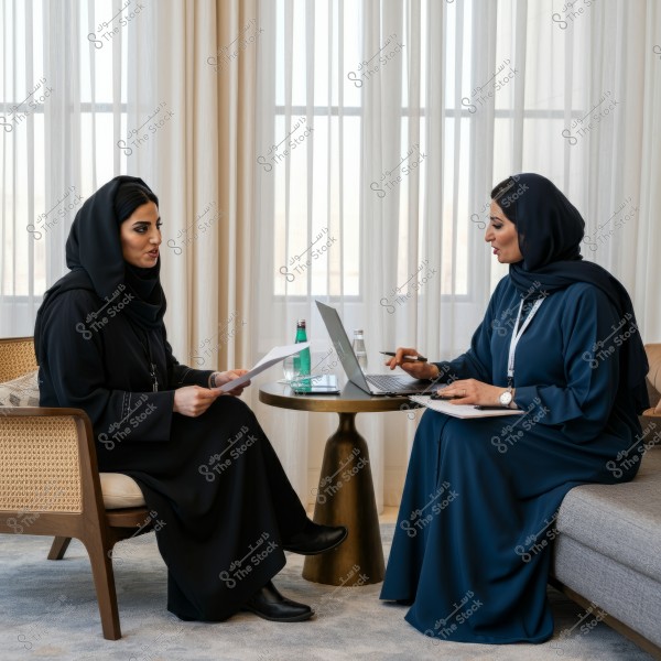Two women wearing traditional Gulf abayas sitting in a comfortable living room. The woman on the left is in a black abaya, holding documents, while the woman on the right is in a dark blue abaya, working on a laptop. There are water bottles and a small table between them, with white curtains covering the windows in the background.