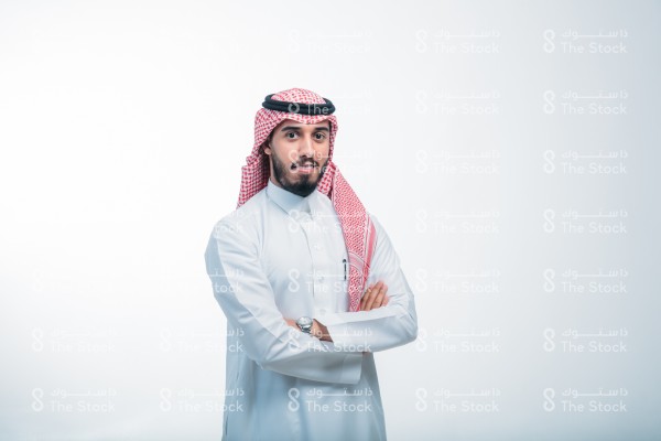 رجل يرتدي الزي الرسمي السعودي ، على خلفية بيضاء ، يقف مبتسماً ، مكتوف اليدين ، صورة شخصية