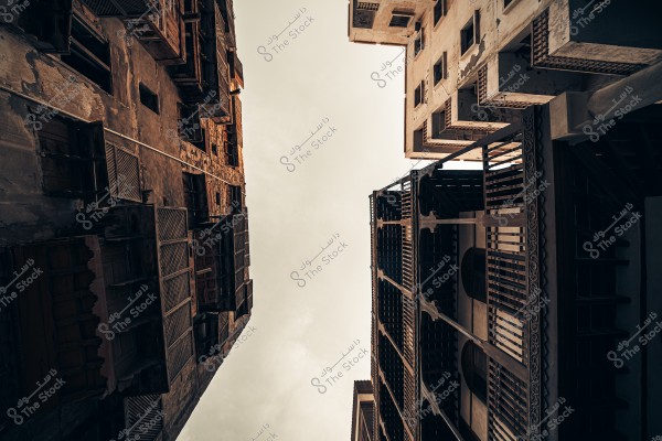 Here’s the translation of your text into English:\r\n\r\n\"A historical image of Jeddah showing a traditional multi-story building at the corner, clearly displaying its unique architectural design with stone and wooden balconies. The surrounding buildings appear slightly worn, giving the area a historical character. The streets are cobblestone, with a wooden chair in front of the central building, and a palm tree visible in the background.\"\r\n\r\nIf you want, I can also make a slightly more **natural, descriptive English version** that reads like a guidebook or historical caption. Do you want me to do that?\r\n