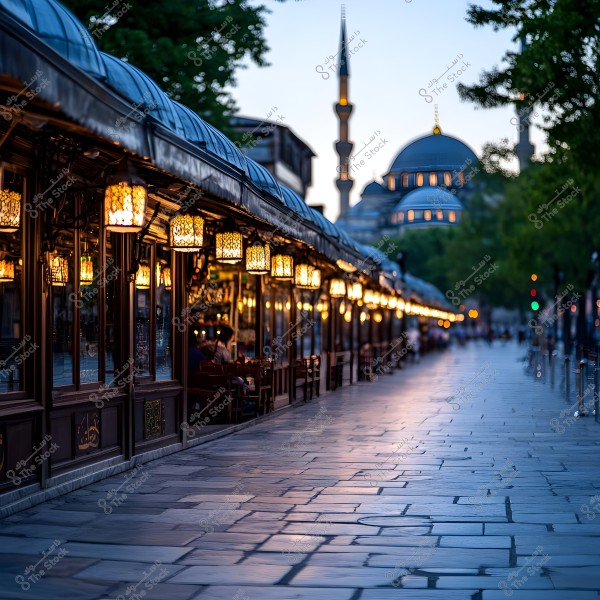 A paved street in the evening is illuminated by glowing lanterns lining the side of an outdoor café. In the background, a mosque with tall minaret towers and a large dome is visible, surrounded by green trees.