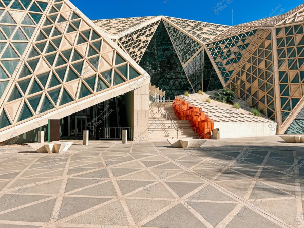Image of a modern building with a complex, angular architectural design. The facade is covered with a pattern of three-dimensional windows arranged geometrically. In the center, there is an entrance and stairs leading to the upper part of the building. Red chairs are lined up on the right side of the stairs, with some plants scattered around the area. The flooring includes tiles with lines forming triangular shapes.