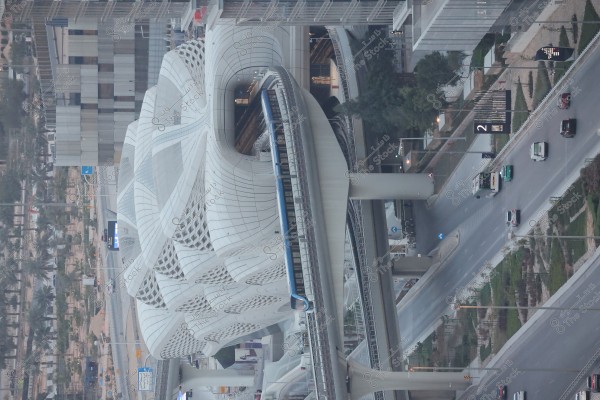An image of a modern train station with an elevated bridge featuring multiple tracks. A blue train is seen approaching the station. The building showcases a complex architectural design with intricate and decorative patterns and large geometric windows. Surrounding the station are buildings and landscaped areas, with cars traveling on the roads below.