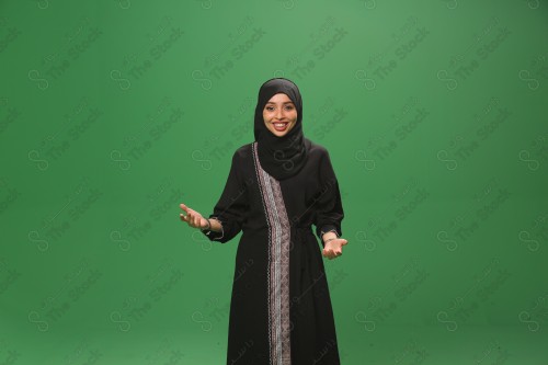 Saudi woman on green background wearing abaya and hijab, making gestures with her hands, extending her hand while smiling, Eid celebrations