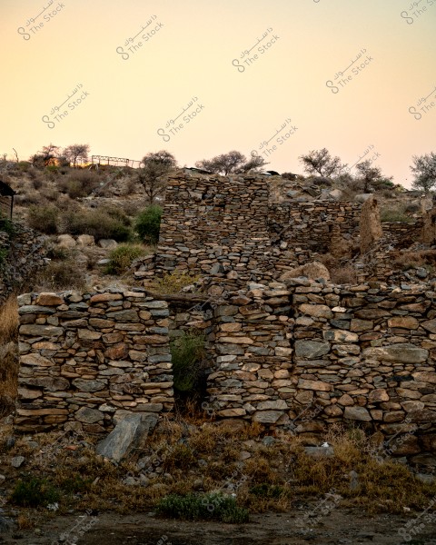 An image of an old, crumbling stone wall in a mountainous area. The wall is constructed from stones of various sizes and shapes. In the background, a few scattered trees are visible on the hillside under a softly colored evening sky.
