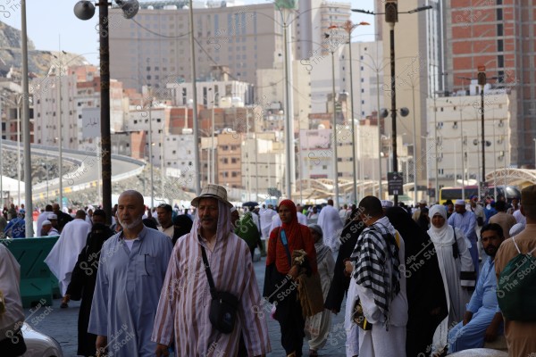 A street scene crowded with pedestrians wearing a variety of clothing in Medina, Saudi Arabia. Tall buildings are visible in the background, representing the city\'s modern architecture. Women are wearing traditional abayas, while men are in traditional attire, with some wearing hats. The street shows a mix of pilgrims and visitors amidst the modern buildings and infrastructure.