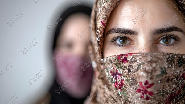 Close-up image of a woman\'s face wearing a brown hijab with floral patterns, with another person in the background wearing a black hijab and a floral cloth covering the face. The eyes are clear and expressive, showing detailed textures in the skin and clothing.