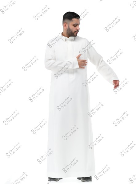 Image of a man wearing a traditional white thobe, looking to the right side with his left hand on his pocket. The man has a short beard and short hair. The background is completely white.