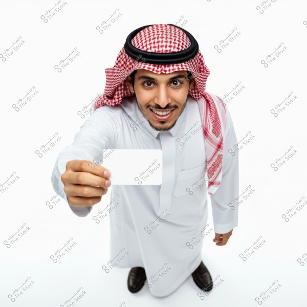 Image of a man wearing traditional Saudi attire, including a white thobe and a red and white checkered ghutra with a black agal. The man is smiling and extending his arm forward, holding a blank white card, and looking at the camera. The background is clean and white.