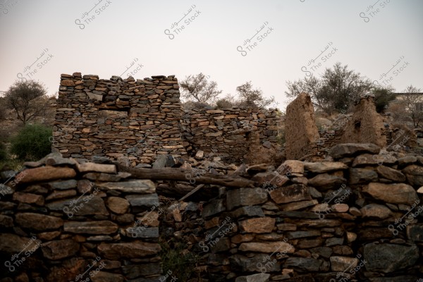 An image showing the remnants of an old stone building in a rural area, constructed from uneven rocks. In the background, some dry trees and a calm daytime horizon can be seen. The structure is roofless, surrounded by crumbling stone walls.