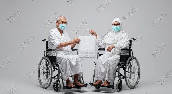 An image of an elderly man and woman seated in wheelchairs against a plain gray background. Both are dressed in white Ihram clothing and wearing medical face masks. They are jointly holding a white shopping bag, symbolizing communication and collaboration. The man is in traditional Ihram attire, while the woman is wearing a hijab and a decorated white abaya.