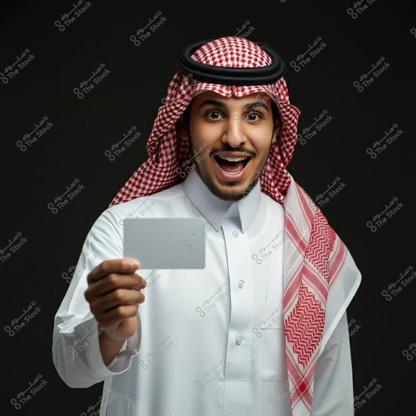 رجل يرتدي ثوبًا أبيض وغترة حمراء وبيضاء, يقف بابتسامة واسعة ويحمل بطاقة بيضاء أمامه. الخلفية سوداء, مما يبرز ابتسامته وملابسه التقليدية.