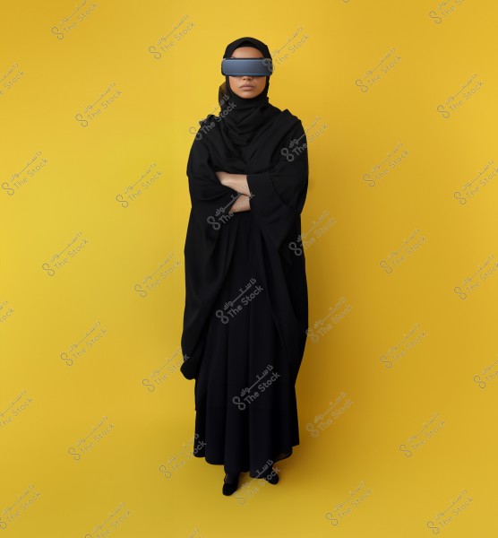 A photo of a woman wearing a black abaya and hijab, standing against a yellow background. She is wearing virtual reality goggles covering her eyes, standing in a frontal pose with her arms crossed, giving a modern and futuristic impression.