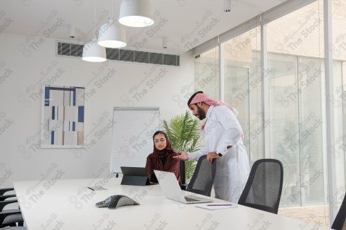 A Saudi man in traditional Saudi dress and a Saudi woman in an abaya discuss working in the meeting room during the day