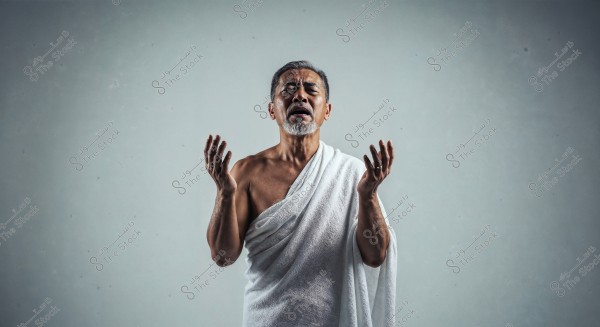 صورة لرجل يقف رافعًا يديه في وضعية دعاء. يرتدي إزارًا أبيض, مما يشير إلى ملابس الإحرام المستخدمة في مناسك الحج أو العمرة. خلفية الصورة بسيطة ومحايدة اللون.