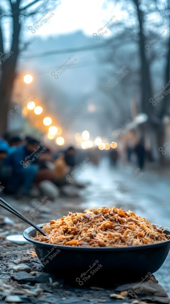 The image shows a large bowl of biryani rice garnished with chicken and nuts like almonds or walnuts, placed on a roadside in a public street. The background is blurred with twinkling night lights and people sitting on the ground, wearing winter clothing.