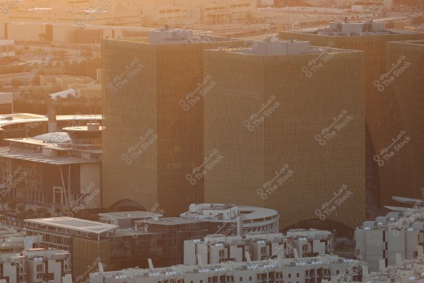 An aerial view of a modern city featuring tall buildings with a contemporary design, appearing golden in color with sunlight reflections. The skyscrapers are surrounded by smaller structures and streets, with an urban backdrop. The sun bathes the city in a warm hue, creating a sunset atmosphere.