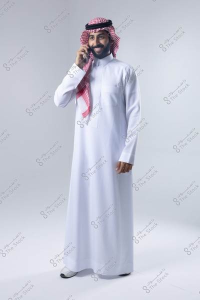 صورة لرجل يرتدي ثوباً أبيض تقليدياً وغترة حمراء وبيضاء مع عقال أسود. يقف في موضع تصوير وهو يتحدث على الهاتف, مبتسماً. يبدو أن الخلفية بيضاء وبسيطة.