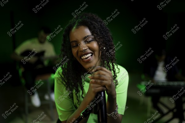 مغنية تبتسم وتمسك بالميكروفون بينما تغني على المسرح, يظهر في الخلفية عازف جيتار وعازف لوحة المفاتيح.