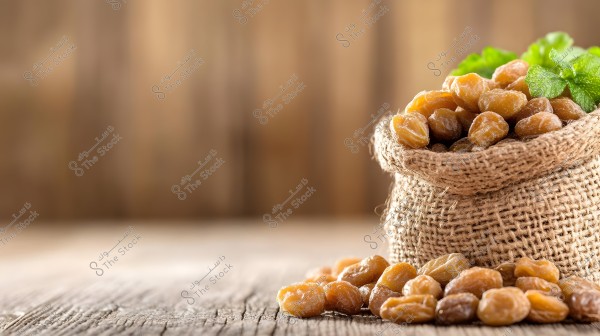 A burlap sack filled with dried chickpeas, placed on a wooden surface. Some chickpeas are scattered outside the sack. The image also features some fresh green leaves protruding from the sack. The background is blurred and appears in light brown.