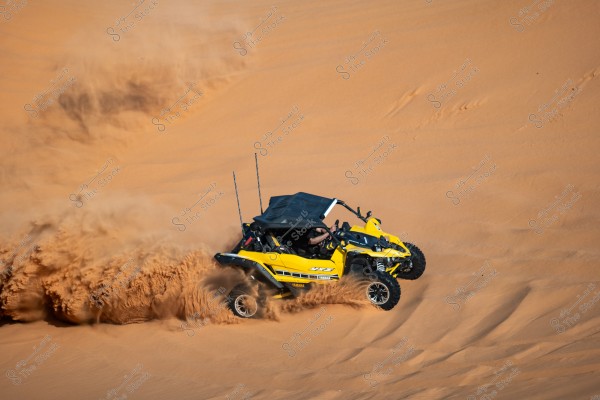 A yellow off-road vehicle speeding across sand dunes in the desert, creating a swirl of sand behind it. The driver is visible in the open cabin of the vehicle, and the surroundings consist of soft, seemingly endless sand.