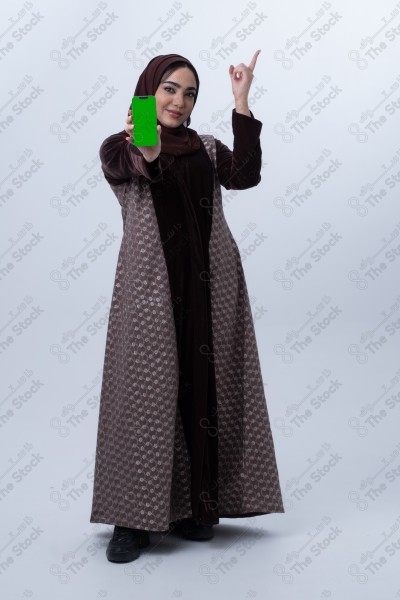 A young Saudi woman wearing an abaya and hijab, holding a green-screen phone