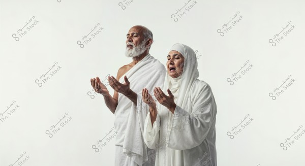 An image of an elderly couple standing side by side in a praying position. The man is wearing a traditional white Ihram garment, and the woman is dressed in a white hijab and abaya. Both have their hands raised and eyes closed, indicating focus and reverence. The background is plain white.