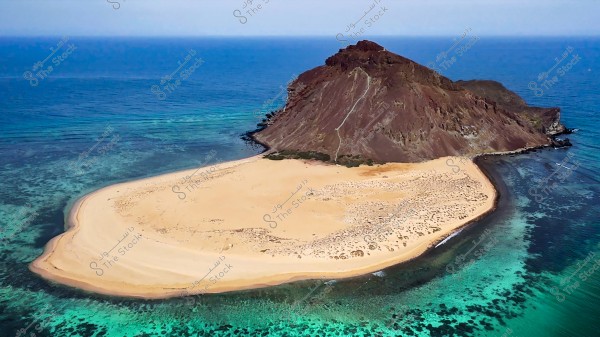 A small island with a sandy beach is shown in the foreground, surrounded by blue and light blue water. A raised brown rocky plateau covers part of the island, and the surrounding waters appear transparent with various shades of the sea.